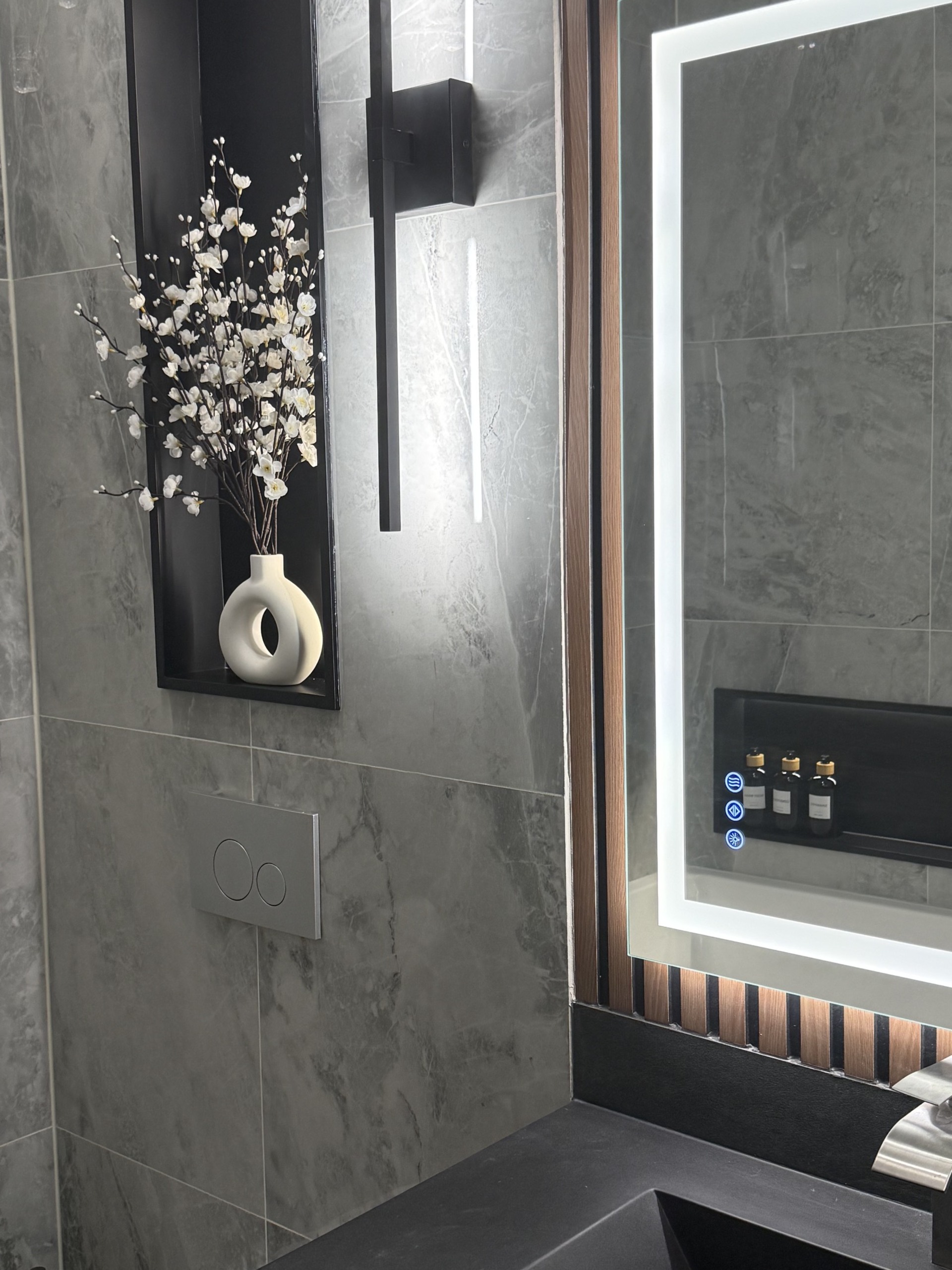 Black wall sconce beside a lit niche with blossoms, next to a backlit vanity mirror
