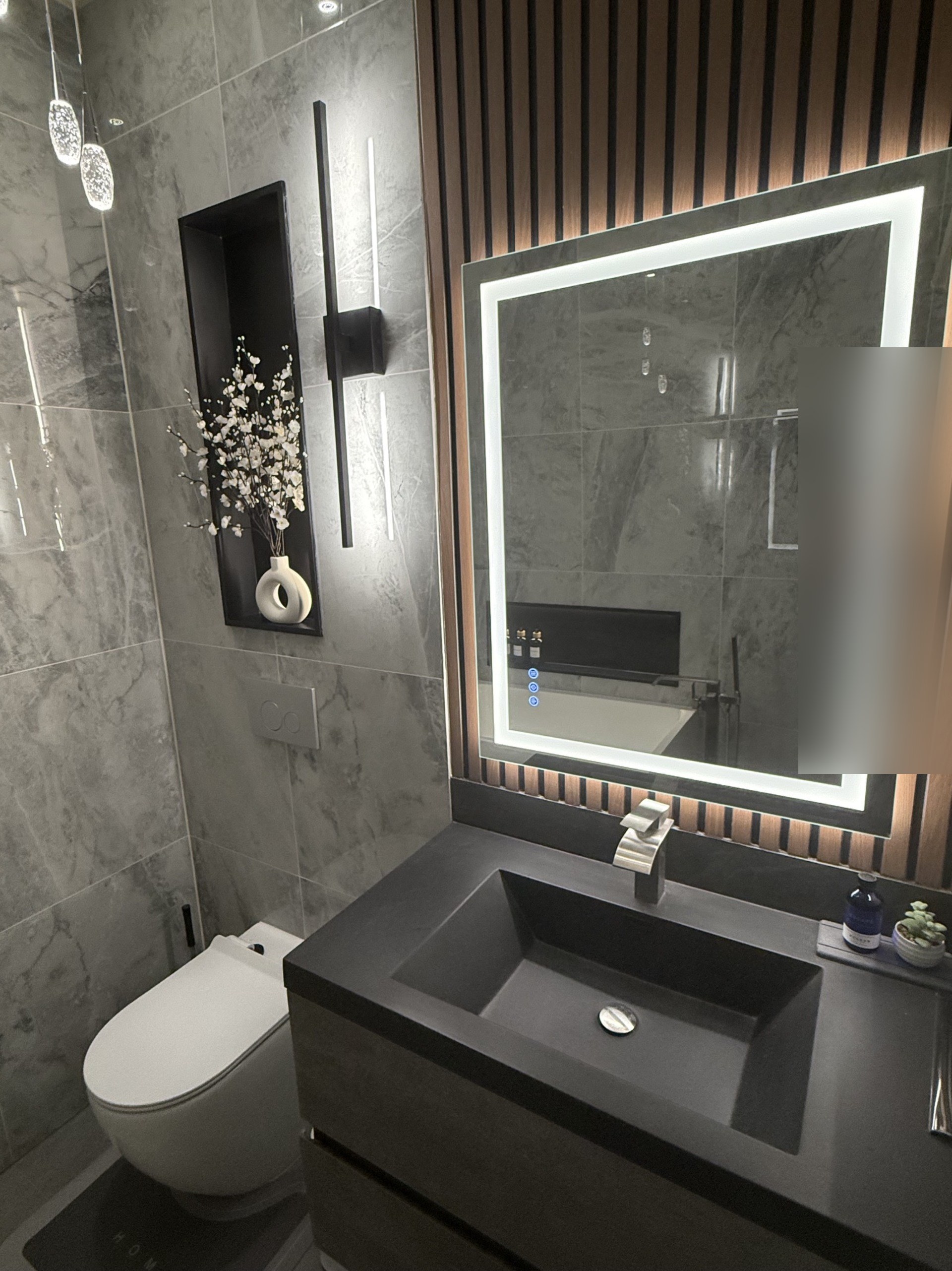 Bathroom with grey marble walls, a black stone vanity, a backlit mirror on a wood-slat wall and a lit niche with blossoms